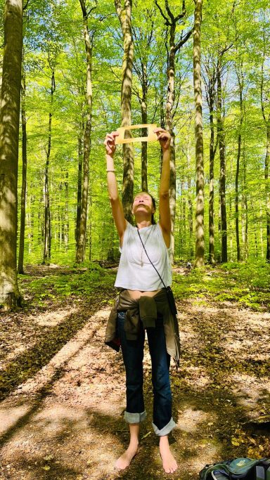 Frau mit hochgehobenen Händen in einem grünen Wald, barfuß und lächelnd.